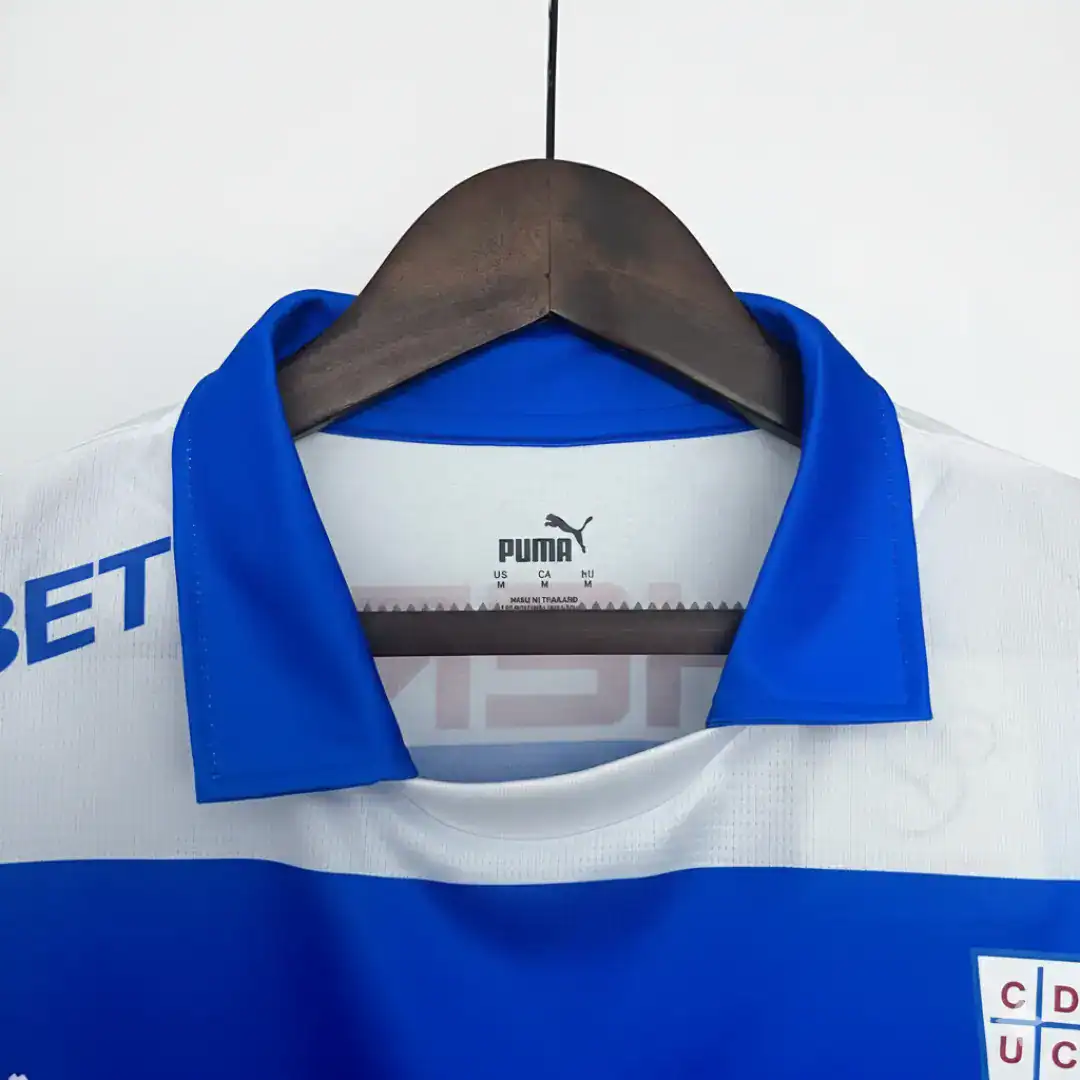 Primer plano de una camiseta de fútbol blanca con un cuello azul vibrante y una franja horizontal azul, mostrando la etiqueta interna de Puma y parte del logotipo del patrocinador 'BET', colgada en una percha de madera.