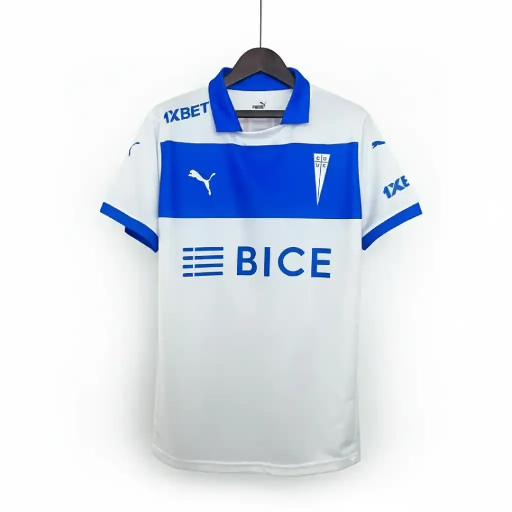 Camiseta de fútbol de Universidad Católica, modelo de local, de color blanco y azul. Presenta una franja azul horizontal en el pecho con el logo de BICE en blanco. El cuello es azul y las mangas tienen detalles azules. Incluye el escudo del club en el lado izquierdo y el logo de Puma en el derecho, ambos en azul. Los patrocinadores 1XBET están visibles en los hombros. La camiseta está colgada sobre un fondo blanco.