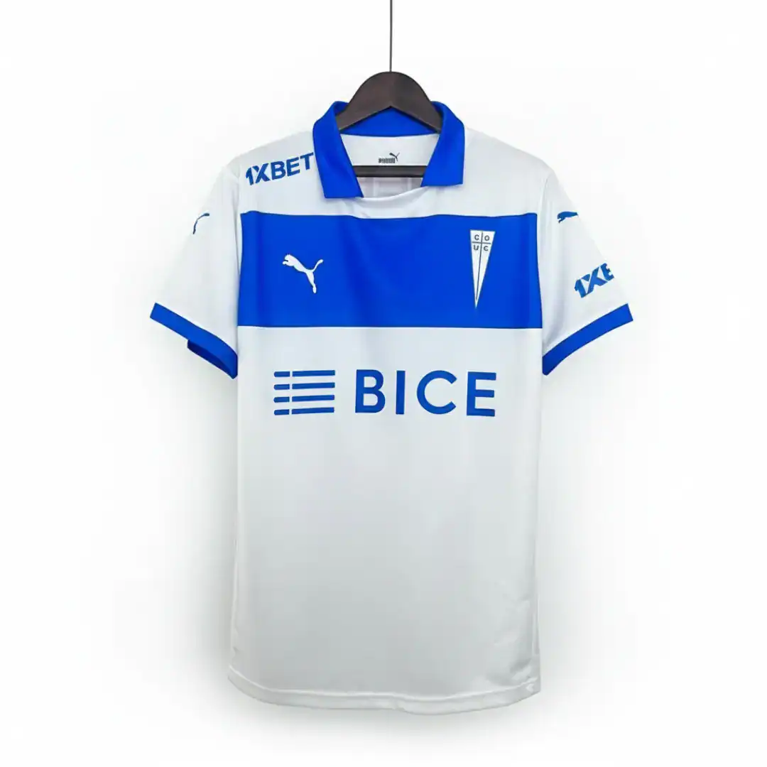 Camiseta de fútbol de Universidad Católica, modelo de local, de color blanco y azul. Presenta una franja azul horizontal en el pecho con el logo de BICE en blanco. El cuello es azul y las mangas tienen detalles azules. Incluye el escudo del club en el lado izquierdo y el logo de Puma en el derecho, ambos en azul. Los patrocinadores 1XBET están visibles en los hombros. La camiseta está colgada sobre un fondo blanco.