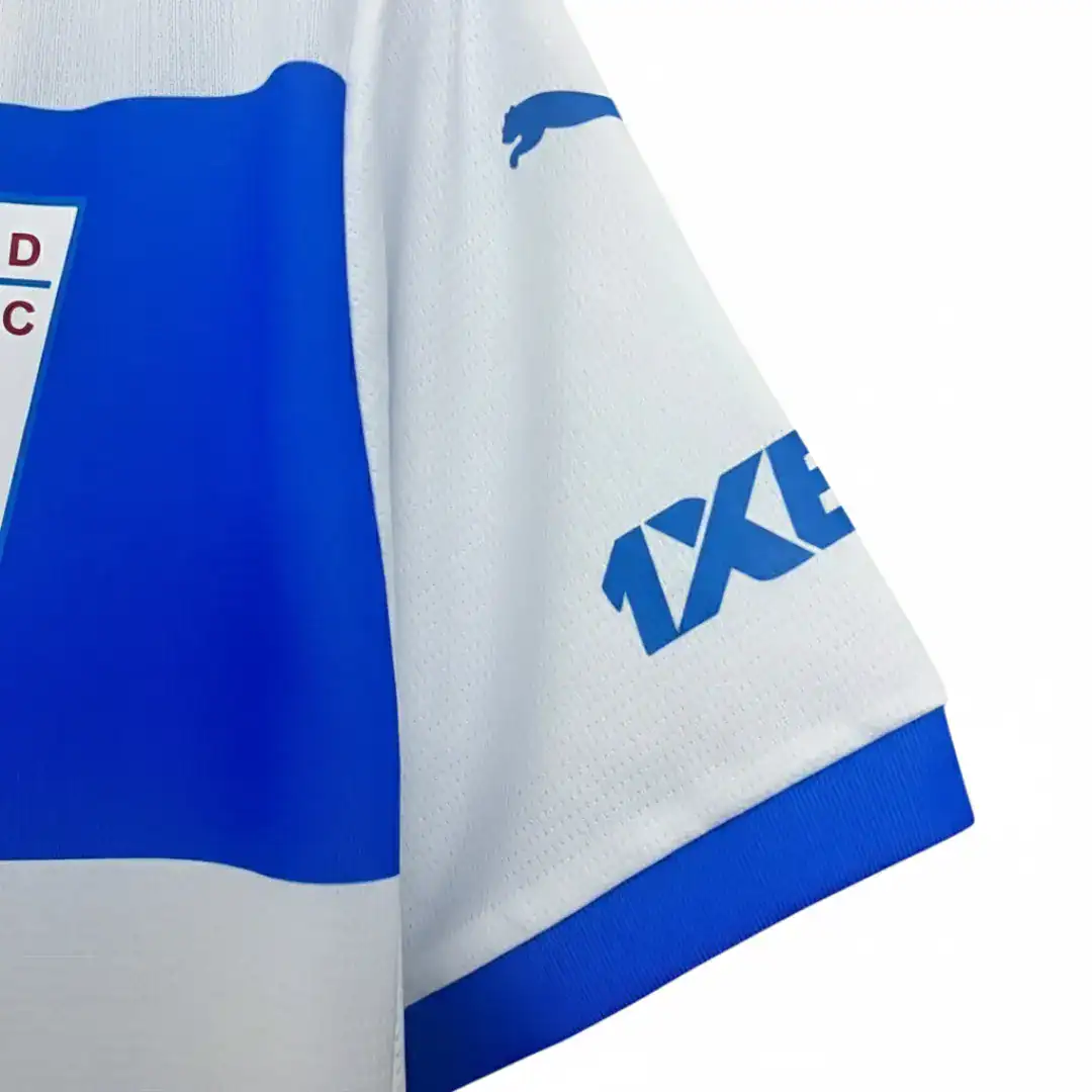 Camiseta de fútbol de Universidad Católica, modelo de local, de color blanco y azul. Presenta una franja azul horizontal en el pecho con el logo de BICE en blanco. El cuello es azul y las mangas tienen detalles azules. Incluye el escudo del club en el lado izquierdo y el logo de Puma en el derecho, ambos en azul. Los patrocinadores 1XBET están visibles en los hombros. La camiseta está colgada sobre un fondo blanco.