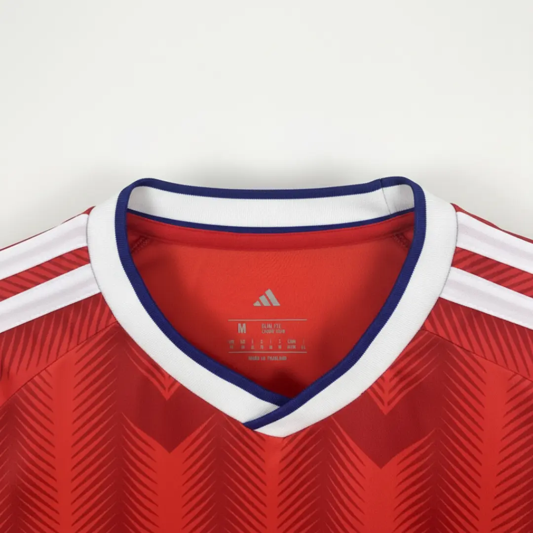 Primer plano de una camiseta deportiva Adidas roja de corte 'Slim Fit' colgada en un gancho, con detalles de cuello blanco y azul, patrón de chevron sutil, y una insignia que celebra 'Clientes Felices' con más de 4.000 reseñas. seleccion chilena 2025