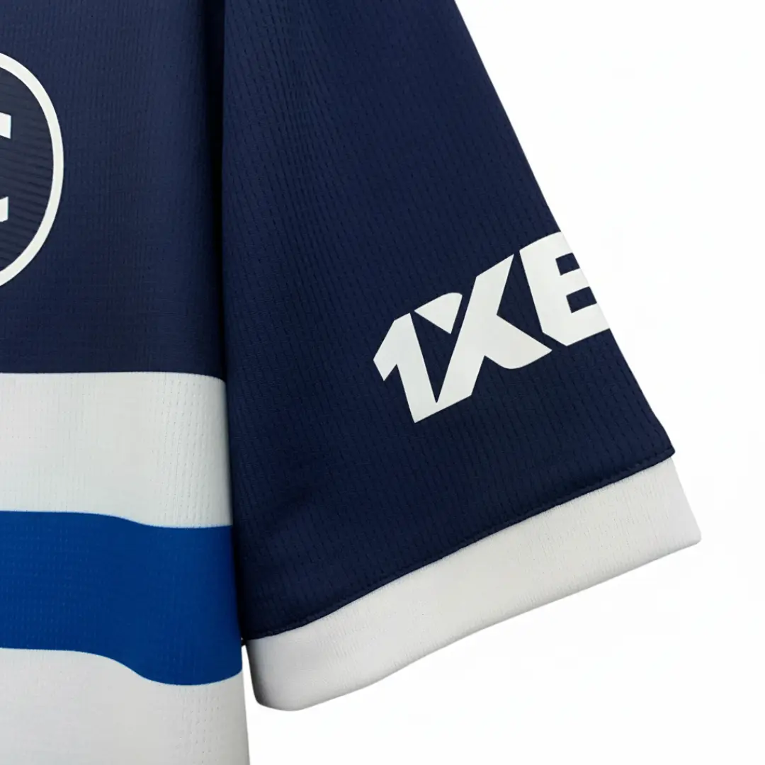 Primer plano de una camiseta deportiva azul marino con un logotipo blanco circular visible parcialmente en el pecho, franjas horizontales blancas y azules, y el logotipo blanco '1XBET' en la manga con un puño blanco.
