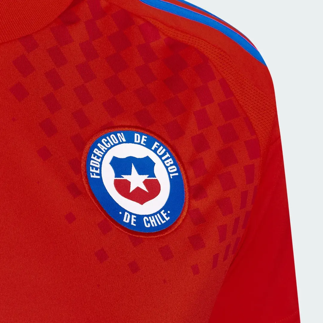 Primer plano de la camiseta de fútbol roja de la selección de Chile, mostrando el emblema redondo de la Federación de Fútbol de Chile con un escudo y una estrella blanca, y franjas azules en el hombro.