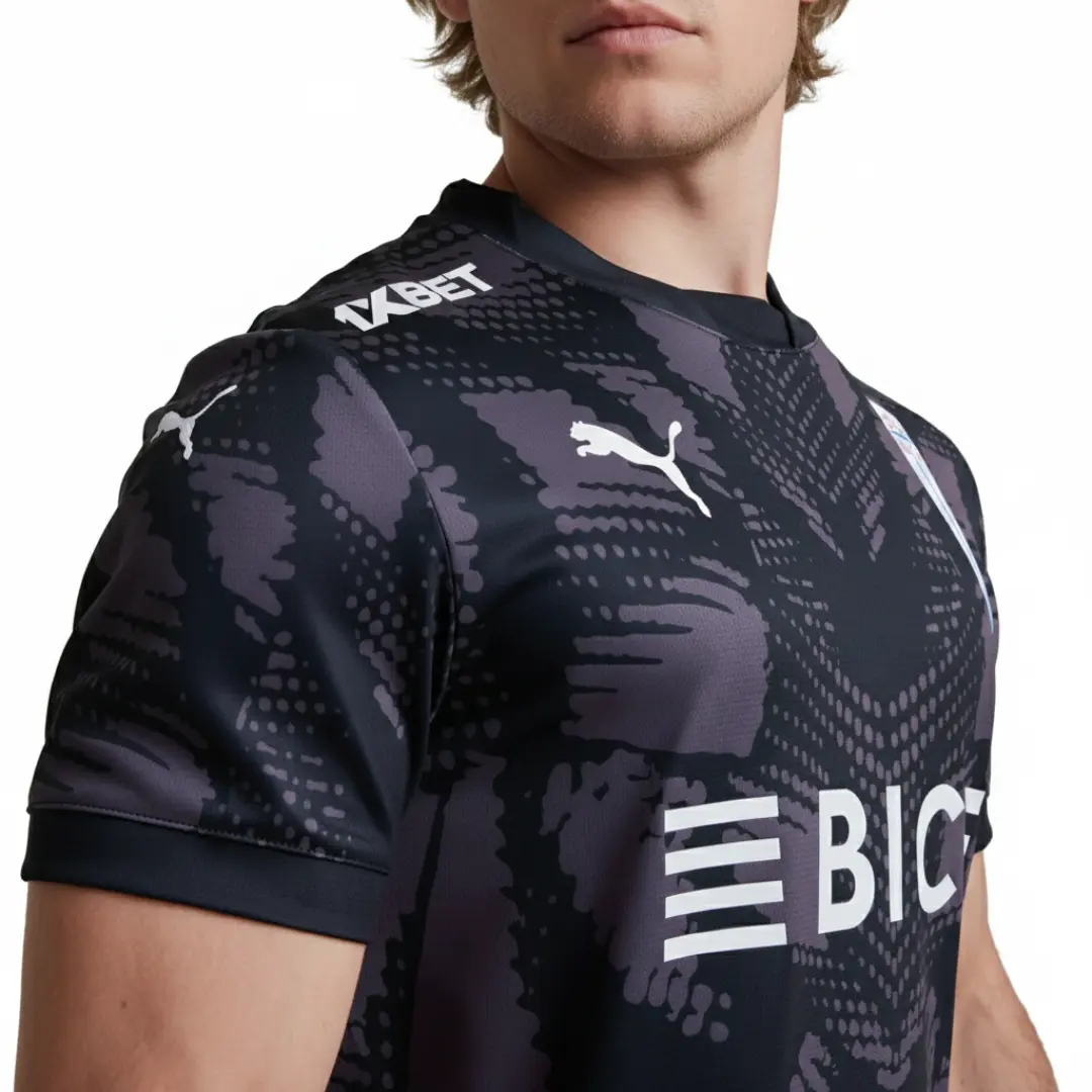 Primer plano de una camiseta deportiva negra con un patrón abstracto oscuro en tonos grises y morados, mostrando logotipos blancos de Puma, 1XBET y parte de BIC en el pecho.