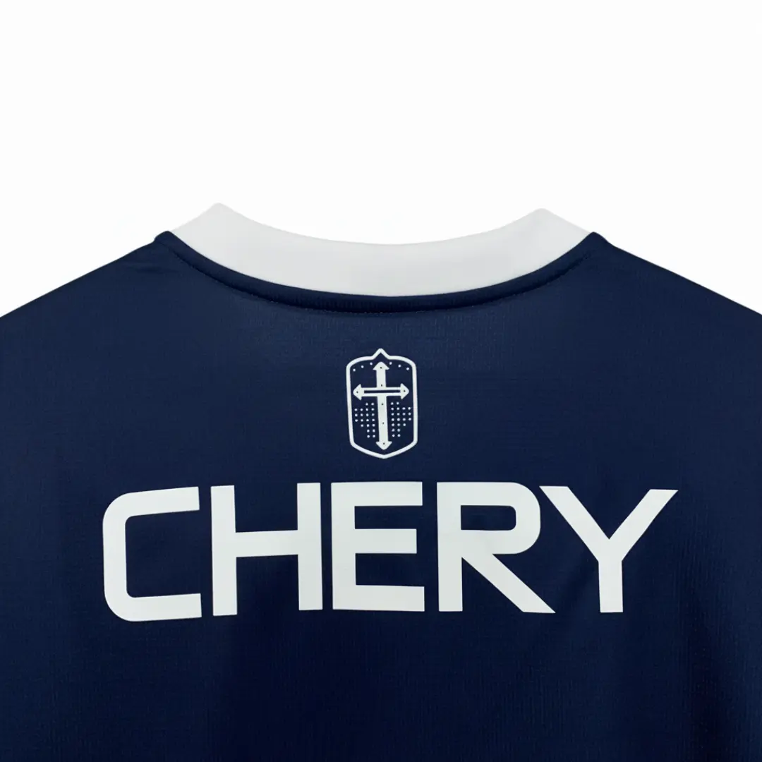 La parte trasera de una camiseta deportiva azul marino con un cuello blanco, un emblema blanco de escudo con una cruz en la parte superior y el nombre 'CHERY' en letras blancas grandes impreso debajo, colgada en una percha de madera.
