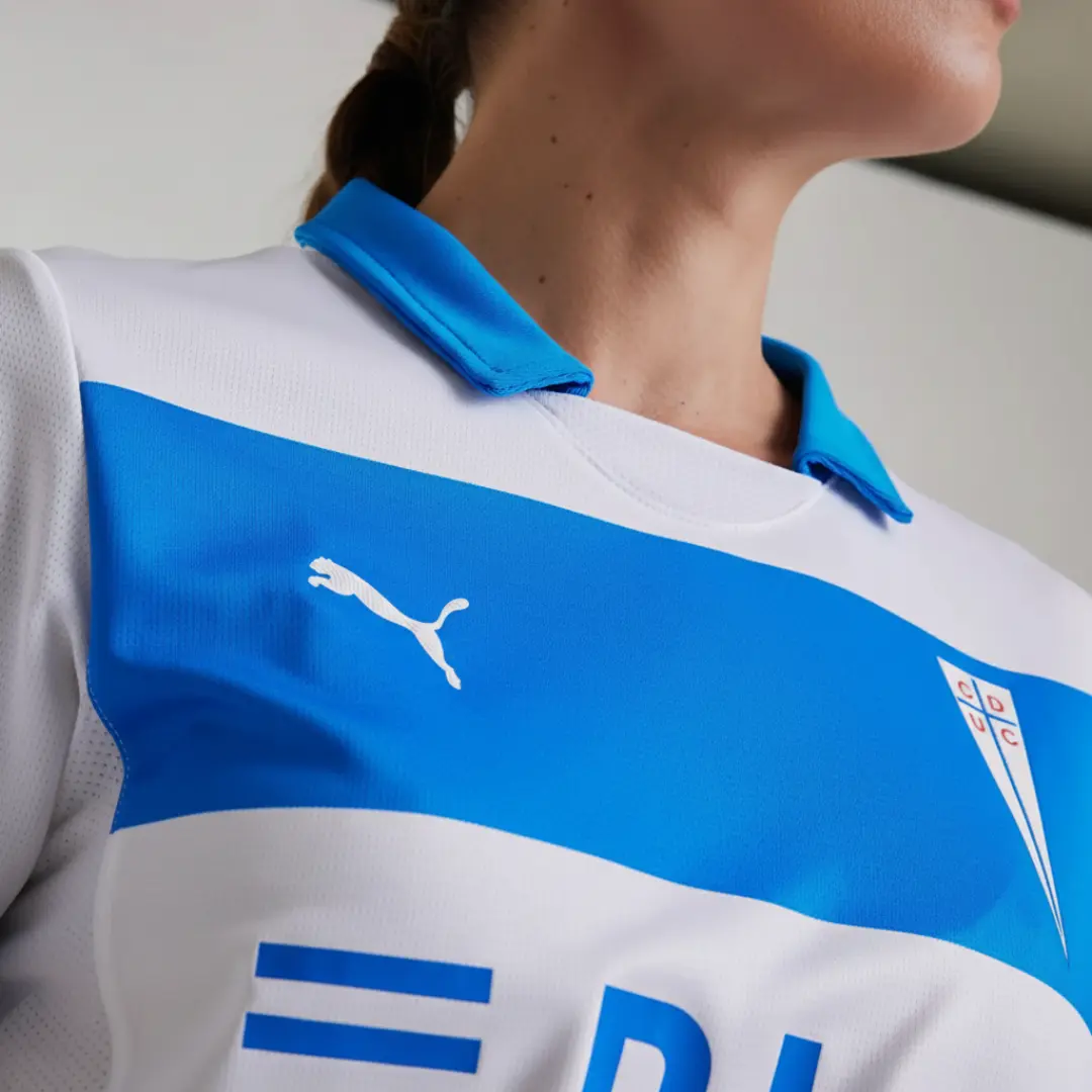 Primer plano de una persona vistiendo una camiseta deportiva blanca con una franja horizontal azul vibrante en el pecho, que muestra el logo blanco de Puma en el lado izquierdo y el escudo del Club Deportivo Universidad Católica (CDUC) en el lado derecho. El cuello de la camiseta es también azul y el tejido es visiblemente transpirable.