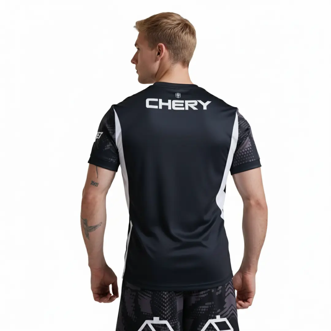 Hombre de espaldas vistiendo una camiseta deportiva oscura con detalles laterales blancos y grises estampados, con el logo blanco de 'CHERY' impreso prominentemente en la espalda. También lleva pantalones cortos estampados oscuros y tiene tatuajes en el brazo izquierdo, mientras mira hacia su derecha con un fondo blanco.