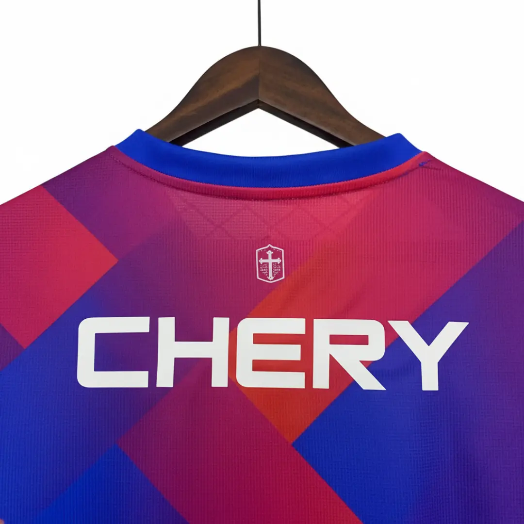 Vista trasera de una camiseta deportiva con un llamativo patrón geométrico en tonos azules, morados y rojos, colgada en una percha de madera. En la parte superior central de la espalda, se observa un pequeño escudo blanco con una cruz, y debajo, el nombre CHERY en grandes letras blancas.