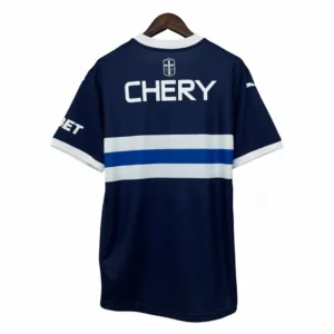Parte trasera de una camiseta de fútbol azul marino oscuro colgada, con cuello y puños blancos. Presenta una franja horizontal blanca y una franja azul más delgada en el centro de la espalda, junto con el logotipo de "CHERY" en blanco y un escudo con una cruz. Se aprecian detalles como el logo de Puma en la manga derecha y parte del texto "BET" en la manga izquierda.