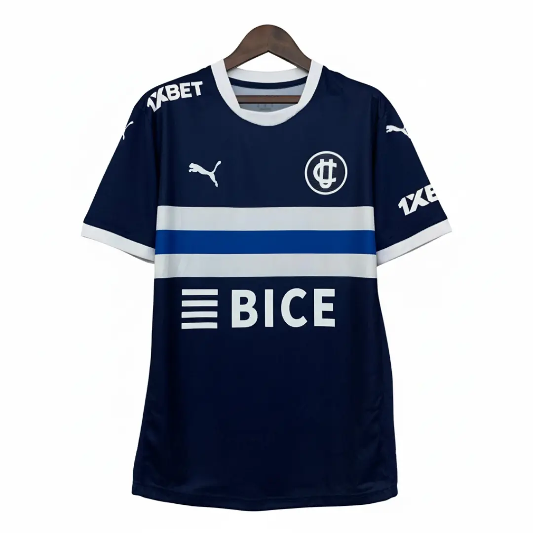 Una camiseta de fútbol del Club Deportivo Universidad Católica de color azul marino, con una franja horizontal blanca y una más delgada azul en el pecho, logos blancos de Puma, el escudo del club, 1XBET en ambas mangas y BICE en la parte inferior central, exhibida sobre un colgador de madera.