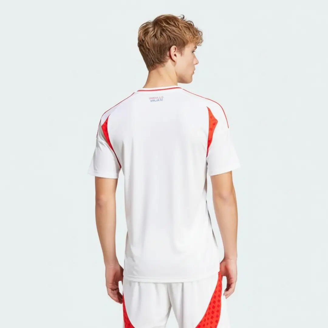 Vista trasera de un hombre vistiendo una camiseta de fútbol blanca con detalles rojos en los hombros y el cuello, y la inscripción 'ORGULLO CHILENO' en la espalda. También lleva pantalones cortos blancos con patrones rojos laterales, posando sobre un fondo claro.