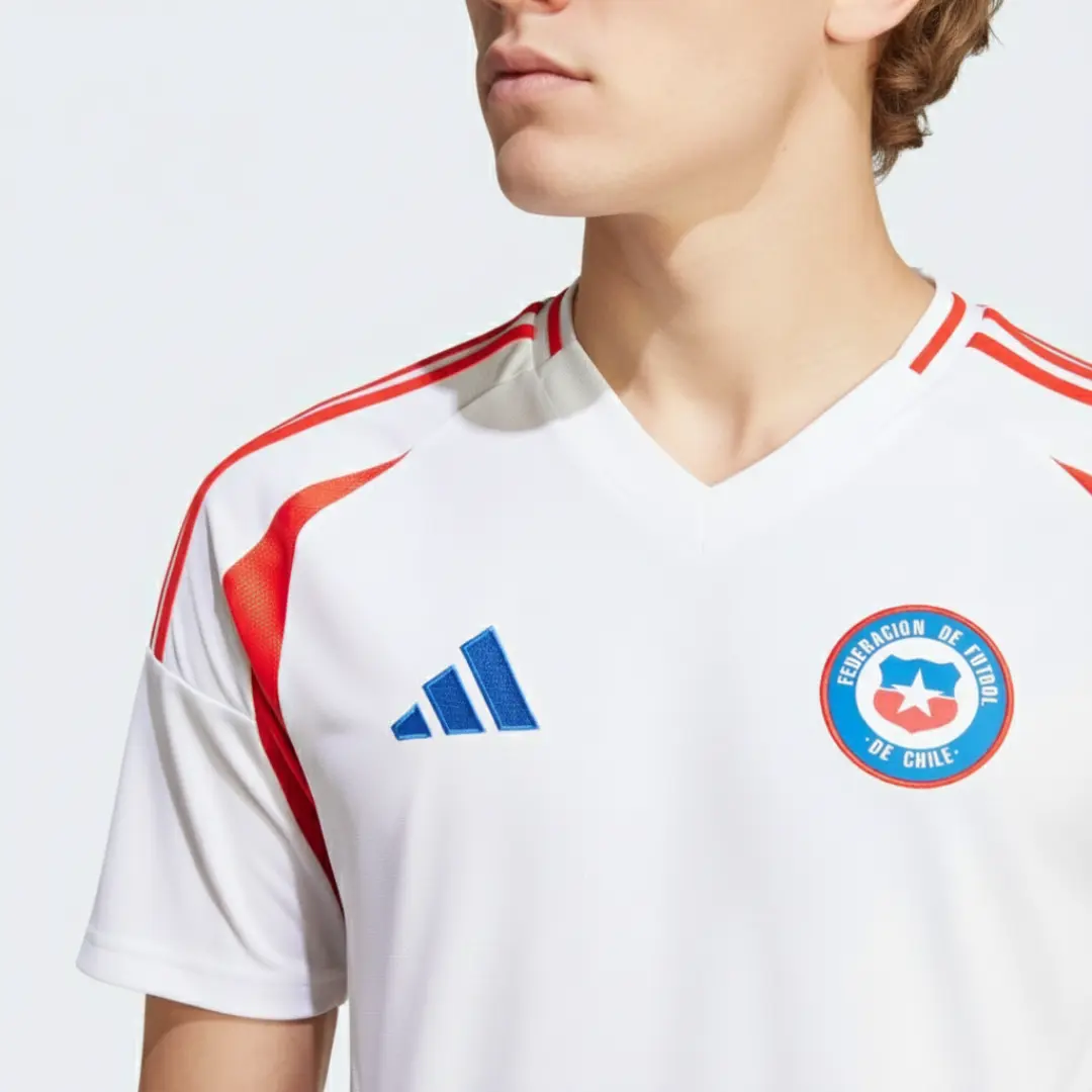 Primer plano de la camiseta de fútbol visitante de la selección de Chile, color blanco, con el logo azul de Adidas en el pecho derecho y el escudo de la Federación de Fútbol de Chile en el pecho izquierdo, ambos sobre un fondo blanco, y las tres rayas rojas icónicas de Adidas adornando los hombros.
