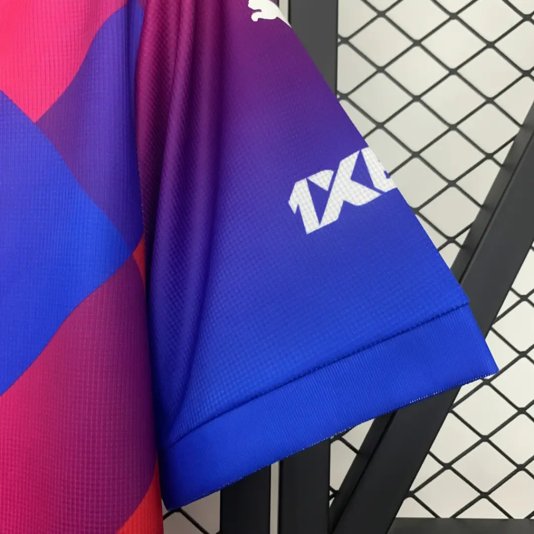 Primer plano de la manga de una camiseta de fútbol con un vibrante degradado de colores morado a azul, mostrando el logo blanco de Puma en el hombro y el patrocinador 1XBET en la manga, con el cuerpo de la camiseta en bloques diagonales rojos y azules.