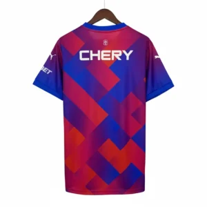 Vista trasera de una camiseta de fútbol con un diseño geométrico vibrante en tonos azul, rojo y morado, mostrando el logotipo blanco del patrocinador 'CHERY' en la parte central, el logo de Puma en la manga derecha y la inscripción 'BET' en la manga izquierda. La camiseta cuelga de una percha de madera sobre un fondo de rejilla metálica.