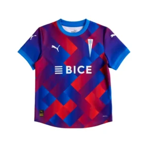 Camiseta de fútbol infantil Puma de Universidad Católica con un diseño vibrante de patrones geométricos en tonos azules, morados y rojos, mostrando el logo del club, el patrocinador BICE y la tecnología Drycell.