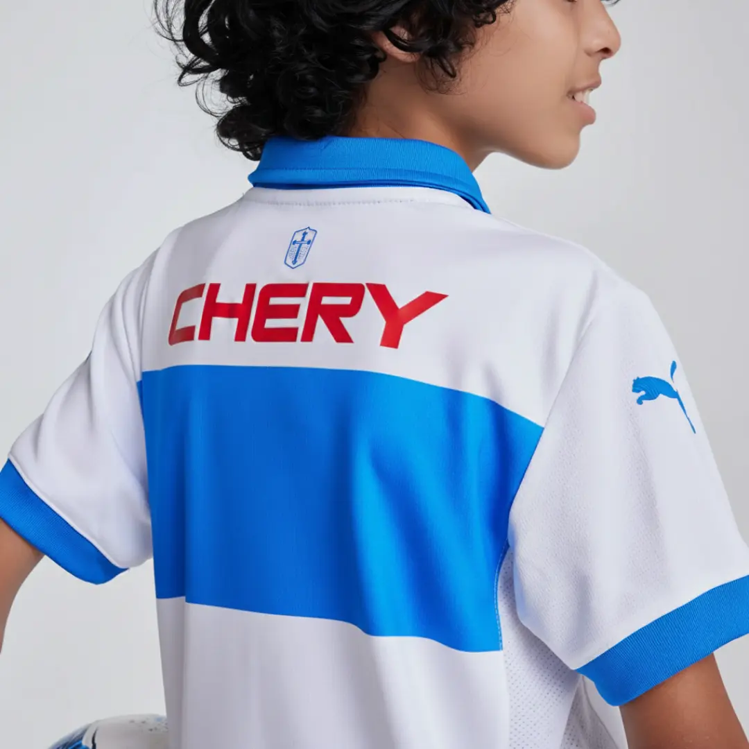 Vista trasera de un niño vistiendo una camiseta de fútbol blanca con franjas horizontales azules, un cuello azul y puños azules, mostrando el logo azul de Puma en la manga derecha, el texto rojo 'CHERY' y un escudo azul en la parte superior central de la espalda.