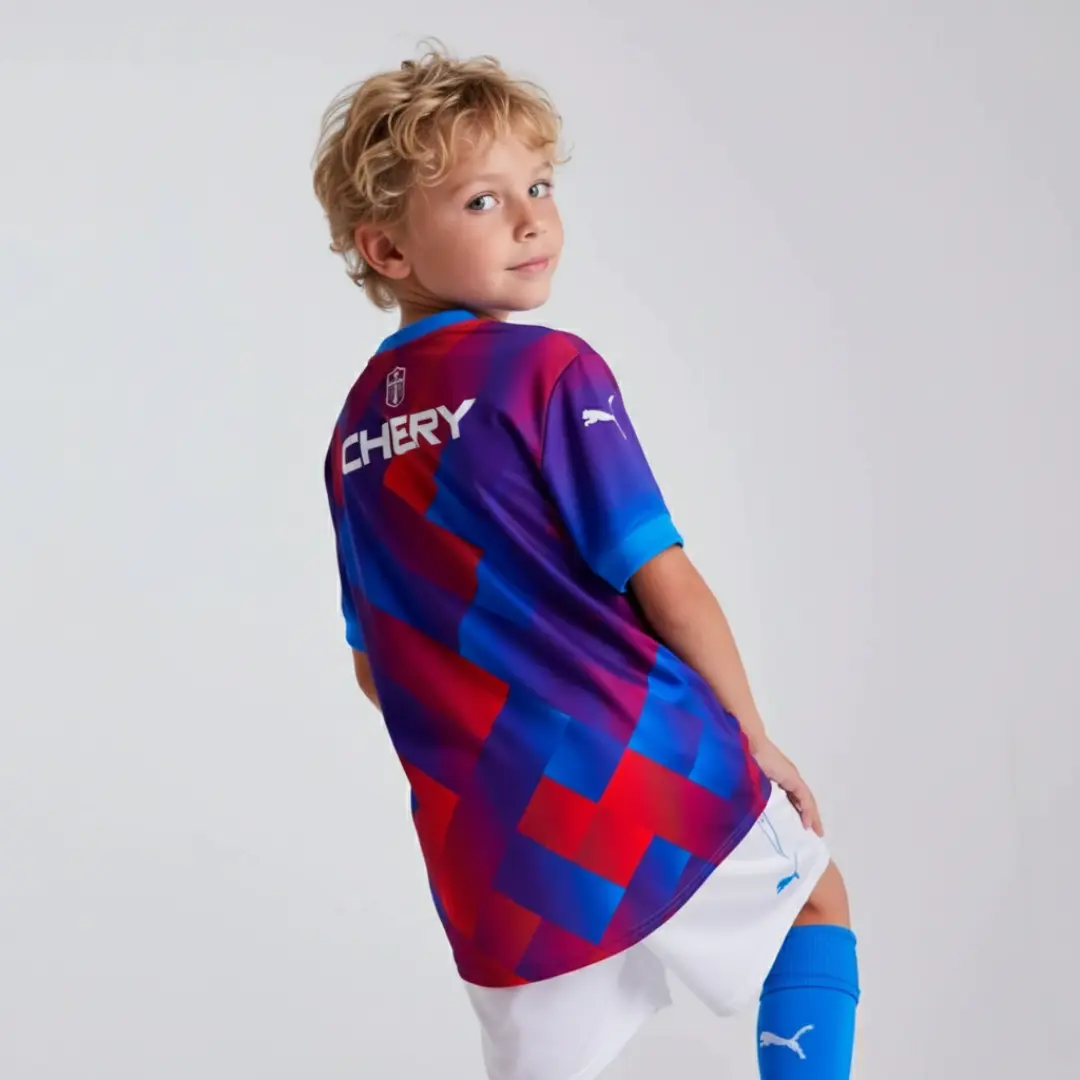 Vista trasera de un niño vistiendo una camiseta de fútbol con un patrón geométrico vibrante en tonos morados, rojos y azules, el logotipo 'CHERY' en blanco en la espalda y el logo de Puma en la manga. El niño también usa pantalones cortos blancos y calcetines azules.