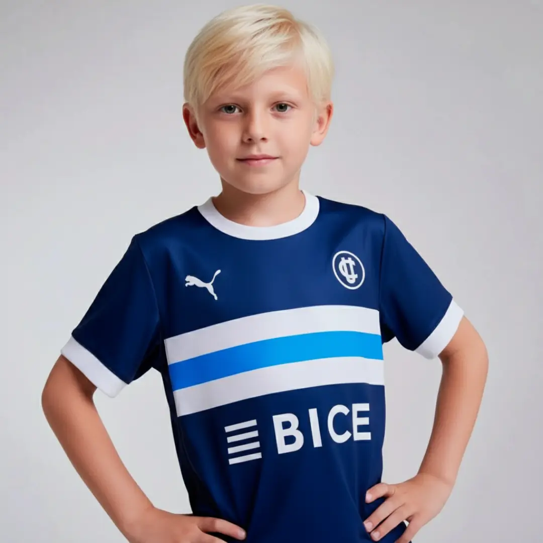 Un niño con cabello rizado oscuro posa sonriendo, vistiendo una camiseta de fútbol azul marino de Universidad Católica con franjas horizontales blancas y celestes en el pecho, el logo de Puma y el patrocinador BICE.