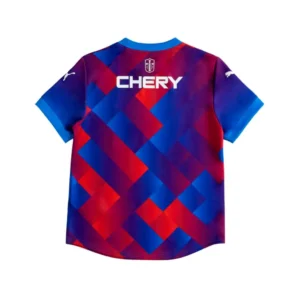 camiseta-futbol-patron-geometrico-rojo-azul-espalda Parte trasera de una camiseta de fútbol con un llamativo diseño geométrico de bloques en tonos rojos y azules. Presenta el logotipo de un escudo con una cruz en la nuca y el patrocinador 'CHERY' en letras blancas grandes. En la manga izquierda se distingue el logo blanco de Puma.
