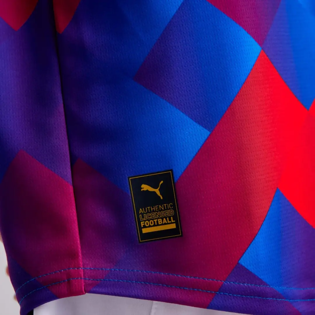 Primer plano de una camiseta de fútbol Puma con un diseño geométrico vibrante en tonos azul, rojo y morado, que incluye la etiqueta de autenticidad negra con el logo dorado de Puma y la inscripción 'AUTHENTIC LICENSED FOOTBALL'.