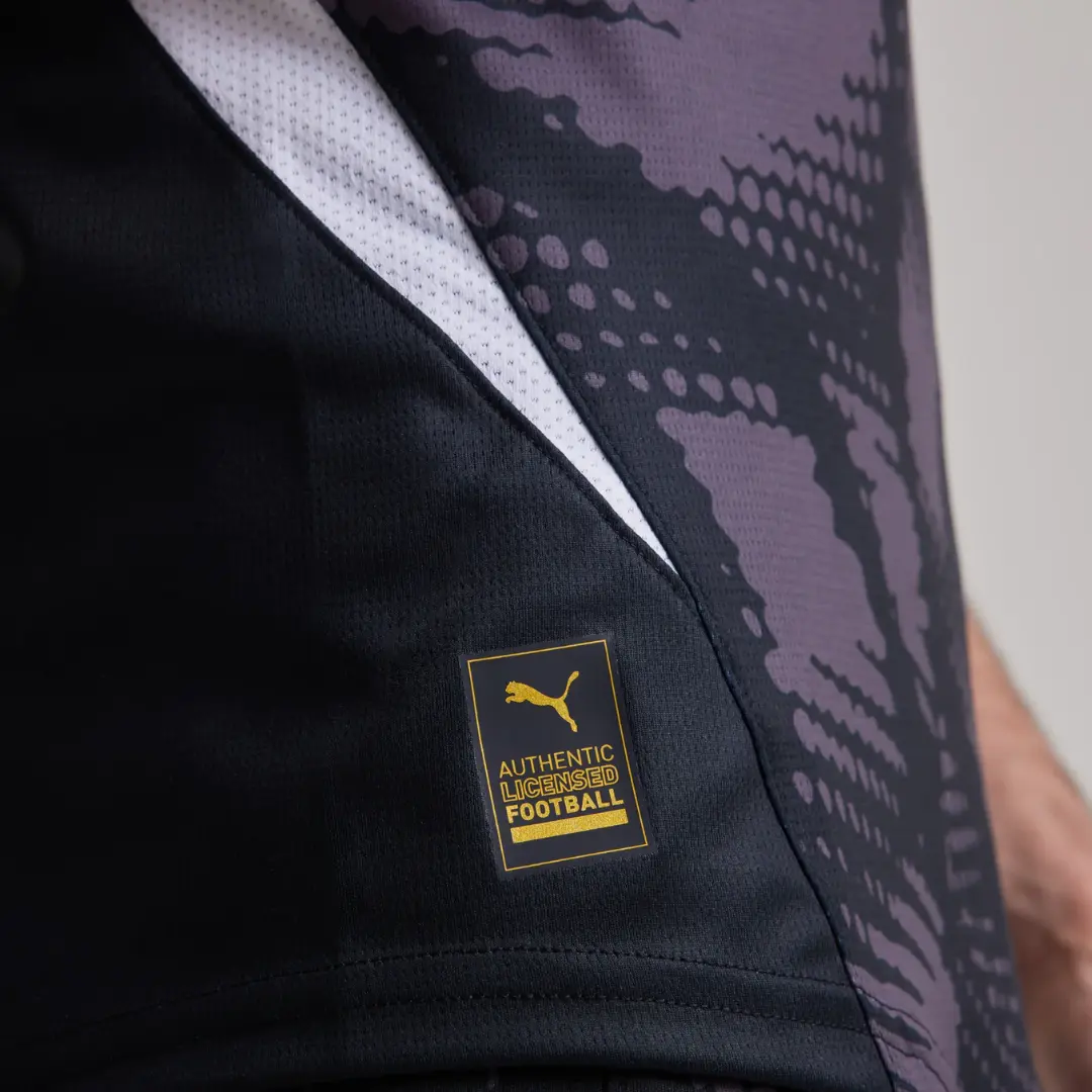 Primer plano de una camiseta de fútbol negra y gris con una franja blanca, destacando una etiqueta dorada de Puma con la inscripción 'AUTHENTIC LICENSED FOOTBALL'.