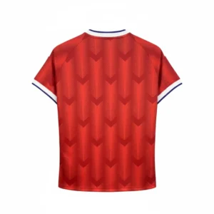 camiseta-futbol-roja-espalda Parte trasera de una camiseta de fútbol roja con un patrón de galones en relieve, cuello blanco y azul, y puños blancos, colgada sobre un fondo de rejilla metálica negra. selccion chilena 2025