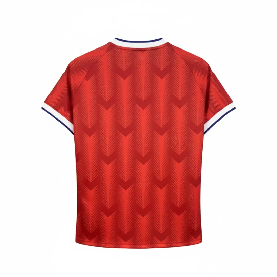 Parte trasera de una camiseta de fútbol roja con un patrón de galones en relieve, cuello blanco y azul, y puños blancos, colgada sobre un fondo de rejilla metálica negra. selccion chilena 2025