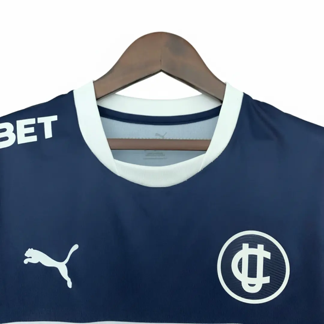 Una camiseta de fútbol azul marino de manga corta, con cuello redondo blanco, colgando de una percha de madera. Presenta el logo blanco de Puma en el lado izquierdo del pecho, el escudo circular blanco del Club Deportivo Universidad Católica con las letras 'UC' en el lado derecho, y el texto blanco 'BET' parcialmente visible en el hombro izquierdo.
