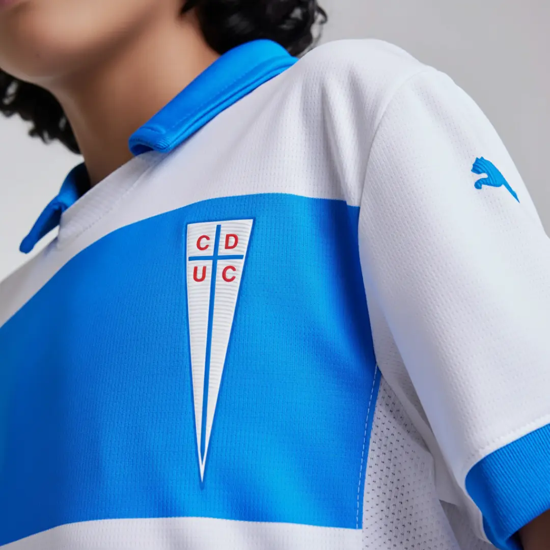Primer plano de una camiseta de fútbol blanca y azul del Club Deportivo Universidad Católica, mostrando el escudo del equipo en el pecho y el logo de Puma en la manga derecha.