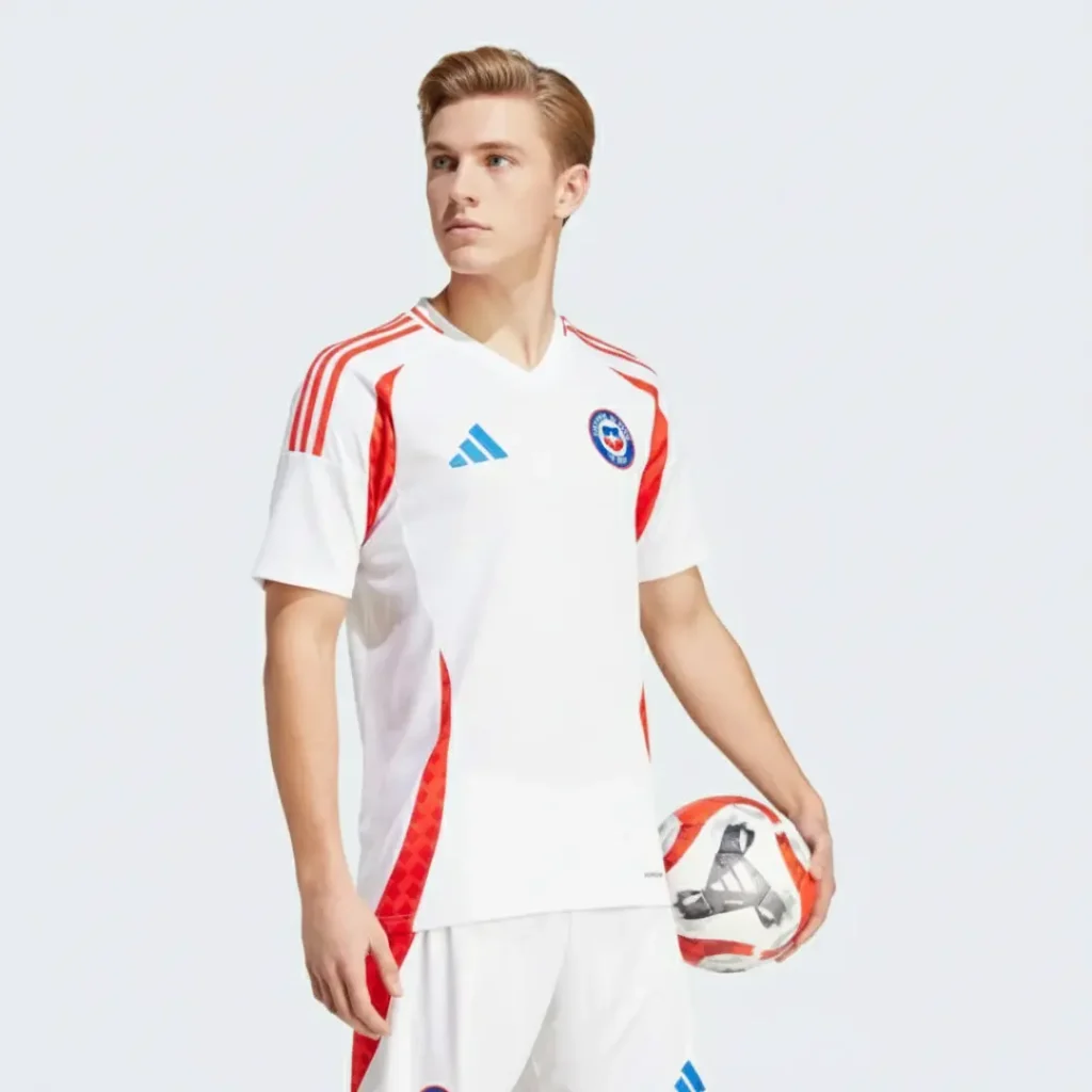 Un modelo masculino viste la camiseta de fútbol de visitante de la selección de Chile, predominantemente blanca con detalles rojos, el logo de Adidas azul y el escudo del equipo chileno, mientras sostiene un balón de fútbol.