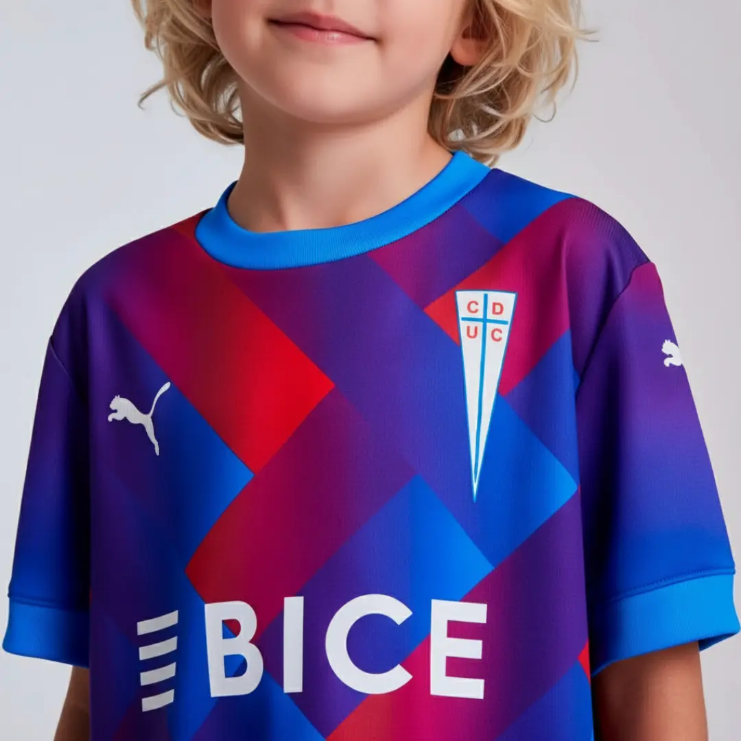 Camiseta de fútbol infantil de Universidad Católica con un llamativo diseño geométrico en tonos azules, púrpuras y rojos, mostrando el escudo del club, el logo de Puma y el patrocinador BICE.