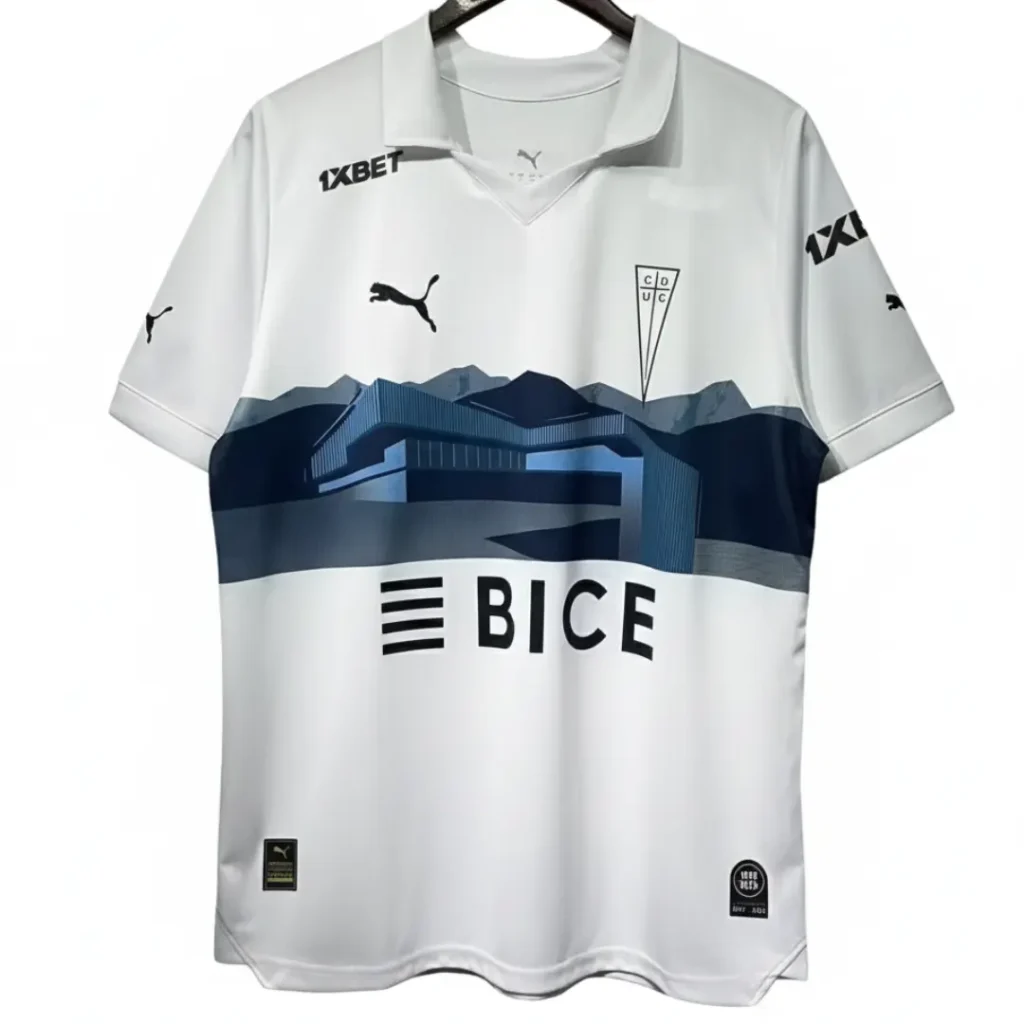 Camiseta oficial de fútbol de la Universidad Católica en color blanco, con un diseño gráfico de montañas y edificios en tonos azules en la parte central, el escudo del club, el logo de Puma y los patrocinadores 1XBET y BICE.