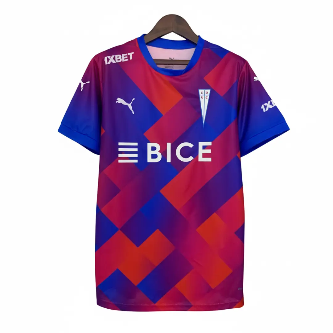Camiseta de fútbol de Universidad Católica, modelo de primera equipación, con un patrón geométrico vibrante de bloques diagonales en tonos azul oscuro, rojo y fucsia, con mangas cortas y cuello azul. Destaca el logo de Puma, el escudo del club CD UC, el patrocinador BICE en el centro y 1XBET en el hombro, todos en blanco, colgando sobre una rejilla metálica.