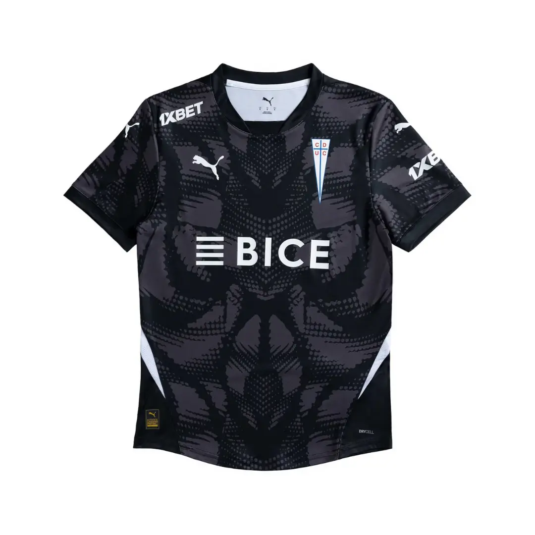 Camiseta de fútbol de Universidad Católica, modelo alternativo, en tonos negros y grises con un patrón abstracto, con los logotipos de Puma, el escudo del club, y los patrocinadores BICE y 1XBET en blanco.
