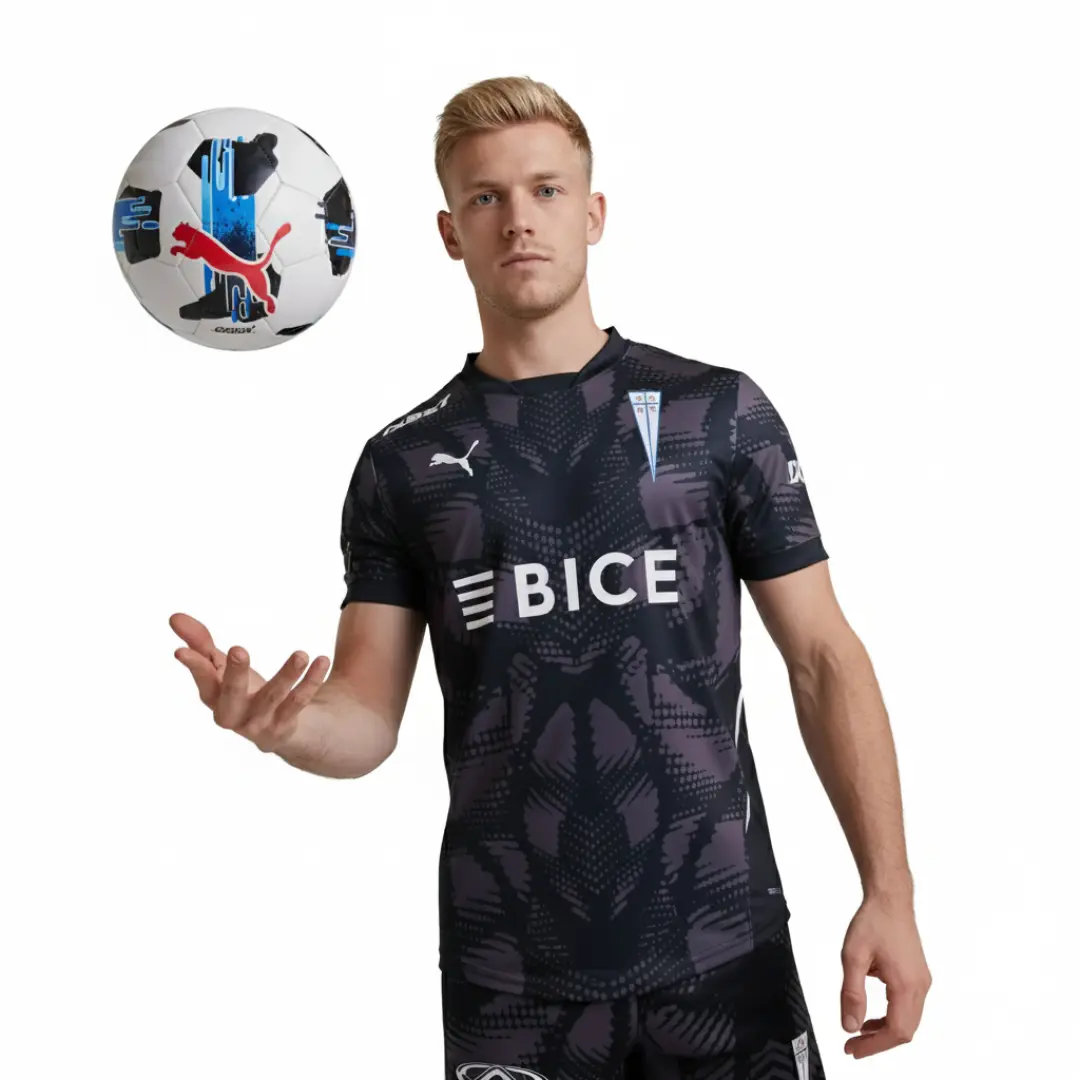 Un modelo masculino con cabello rizado castaño posa vistiendo la camiseta de fútbol visitante de Universidad Católica, de color negro con un patrón gris y detalles del club y patrocinadores como BICE, mientras una pelota de fútbol Puma blanca con detalles azules flota en el aire a su lado.