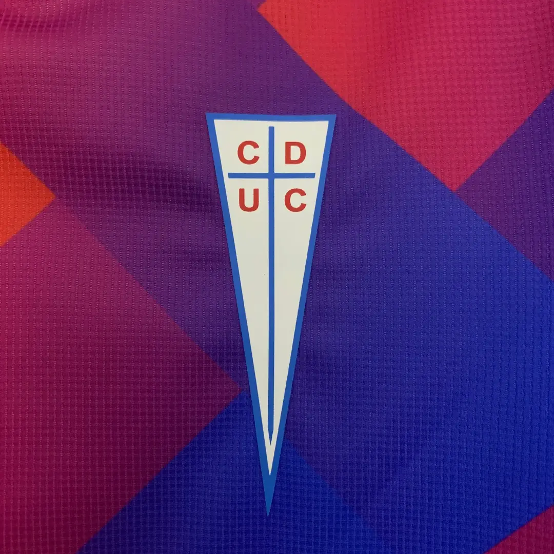 Primer plano del escudo del Club Deportivo Universidad Católica (CDUC) con letras rojas sobre un fondo blanco y bordes azules, centrado en una camiseta de fútbol con un vibrante patrón geométrico en tonos de azul, morado y rojo.