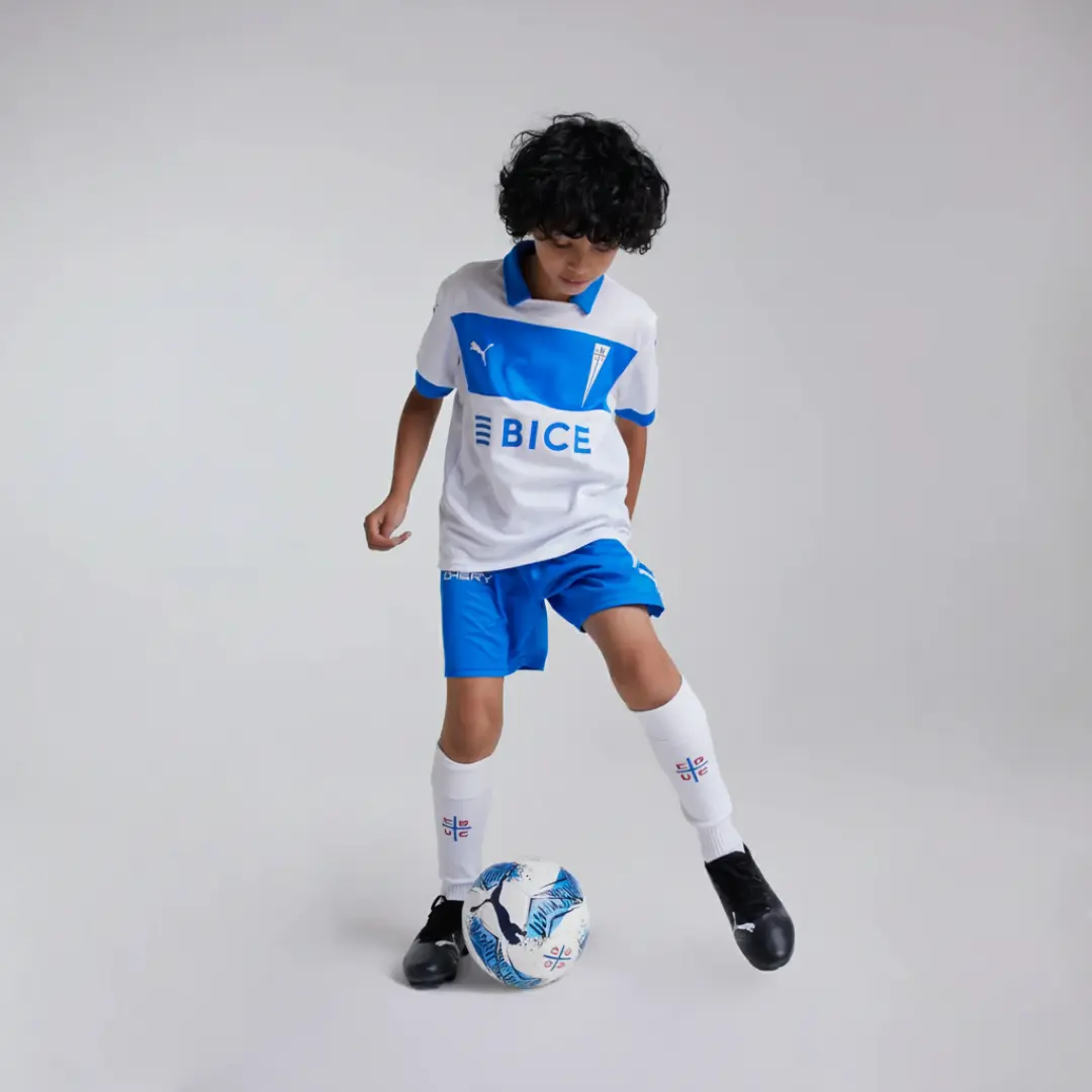 Un niño con el uniforme de fútbol blanco y azul de Puma y BICE, compuesto por camiseta, pantalones cortos y medias, driblando un balón con el pie izquierdo sobre un fondo blanco.