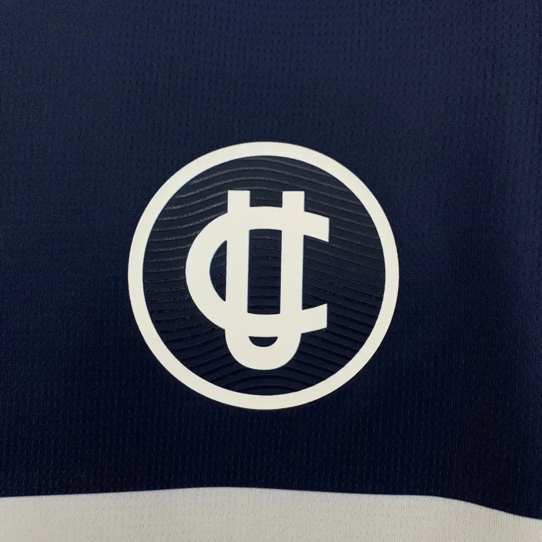 Primer plano de un logo circular blanco con las letras "UC" estilizadas en relieve sobre una camiseta deportiva azul marino texturizada, con una franja blanca en la parte inferior.