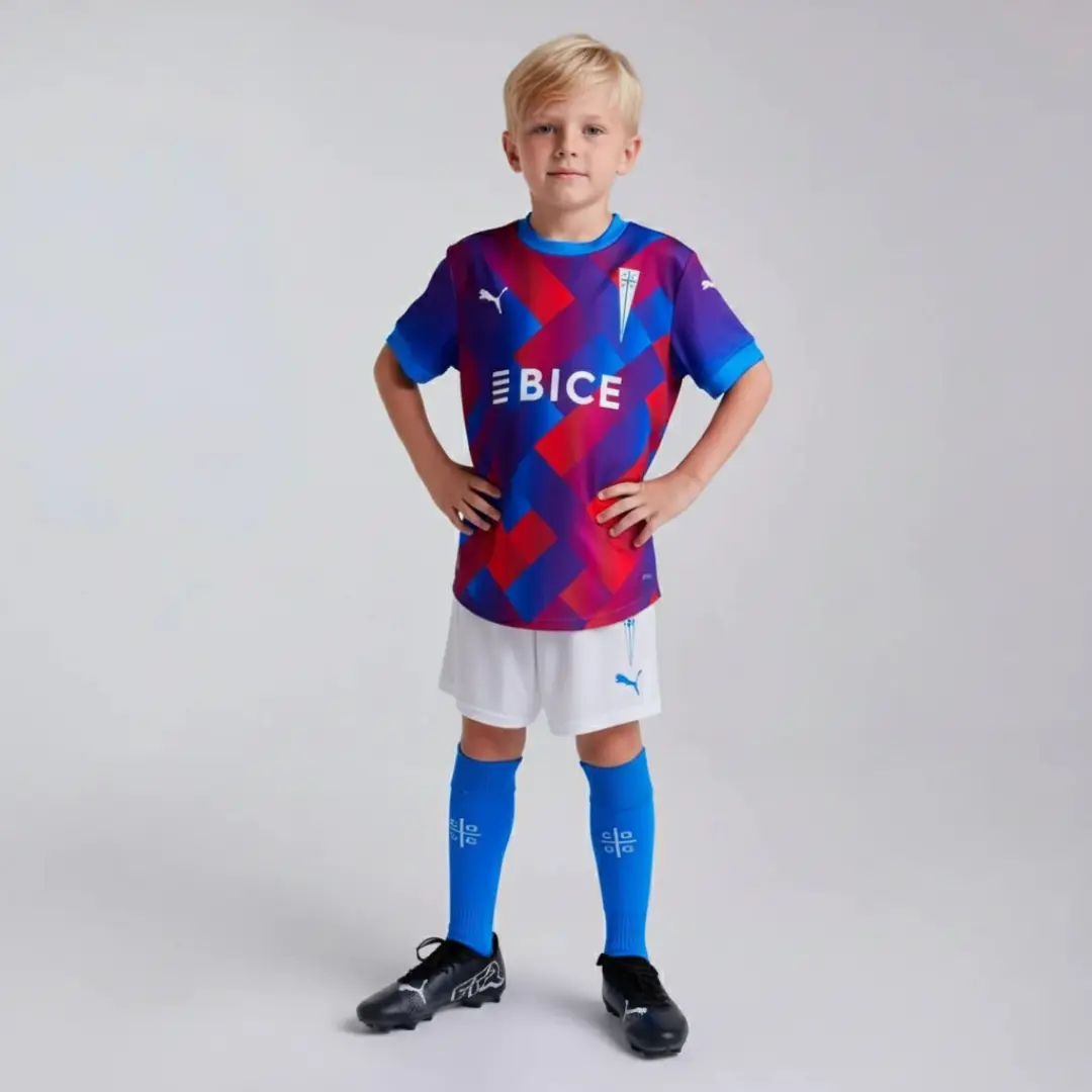Un niño joven con cabello rizado posa con las manos en las caderas, vistiendo un uniforme de fútbol vibrante. La camiseta es de manga corta con un patrón geométrico en tonos azul, rojo y morado, con el logo 'BICE' y un escudo. Complementa su atuendo con pantalones cortos blancos, calcetines azules con un logo blanco y botines de fútbol negros.