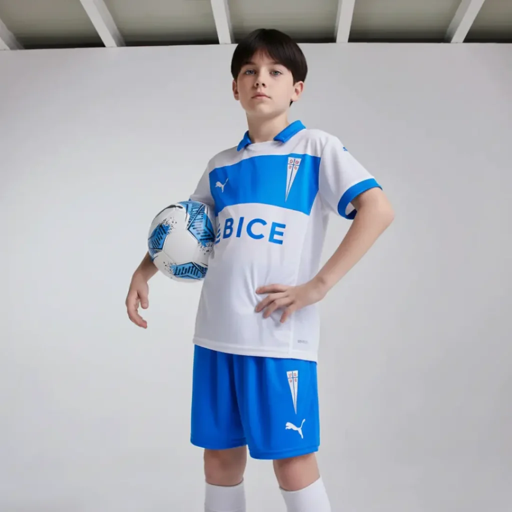 Retrato de un niño con cabello rizado oscuro, vistiendo el uniforme de fútbol blanco y azul de la Universidad Católica, que incluye una camiseta con la franja de BICE y el escudo del club, y shorts a juego, mientras sostiene un balón de fútbol blanco y azul bajo el brazo.