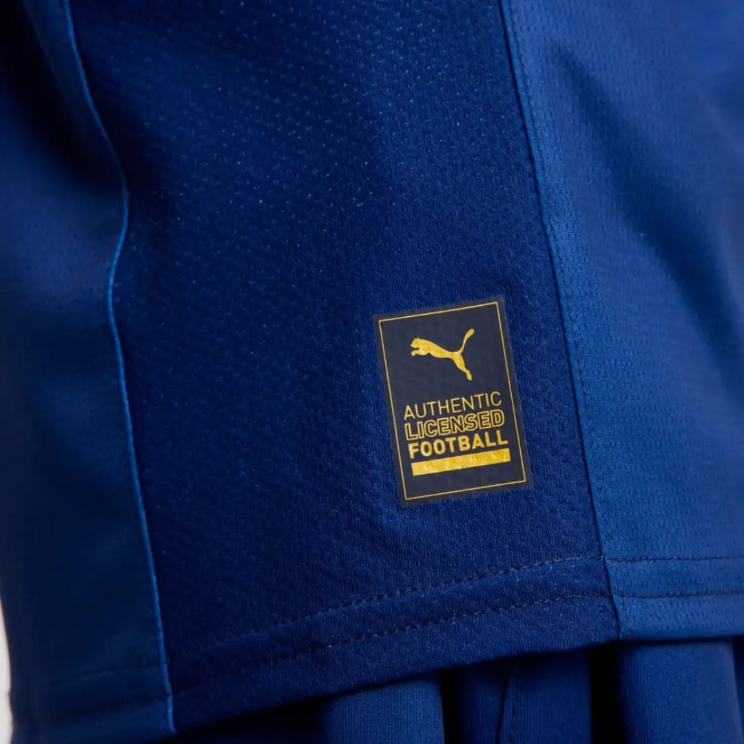 Detalle de una tela deportiva azul con textura transpirable, mostrando un parche dorado de 'AUTHENTIC LICENSED FOOTBALL' y el logo de Puma, indicando autenticidad del producto.