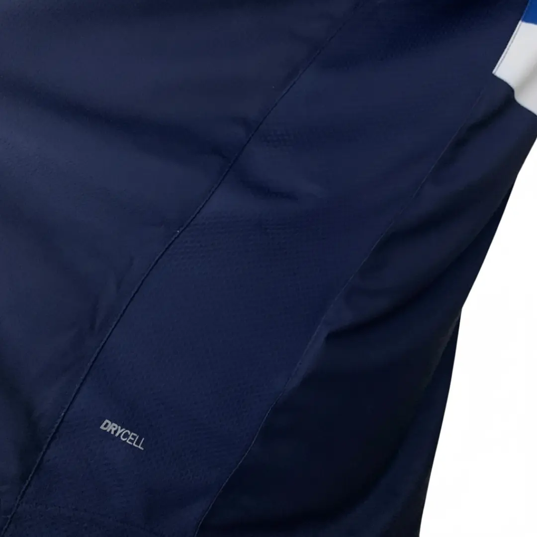 Primer plano de una prenda deportiva azul marino con tecnología DryCELL, mostrando una costura diagonal que divide secciones de tela lisa y texturizada, con el logotipo brillante DRYCELL visible en la parte inferior izquierda y un detalle de franja blanca en la esquina superior derecha sobre un fondo de rejilla metálica.