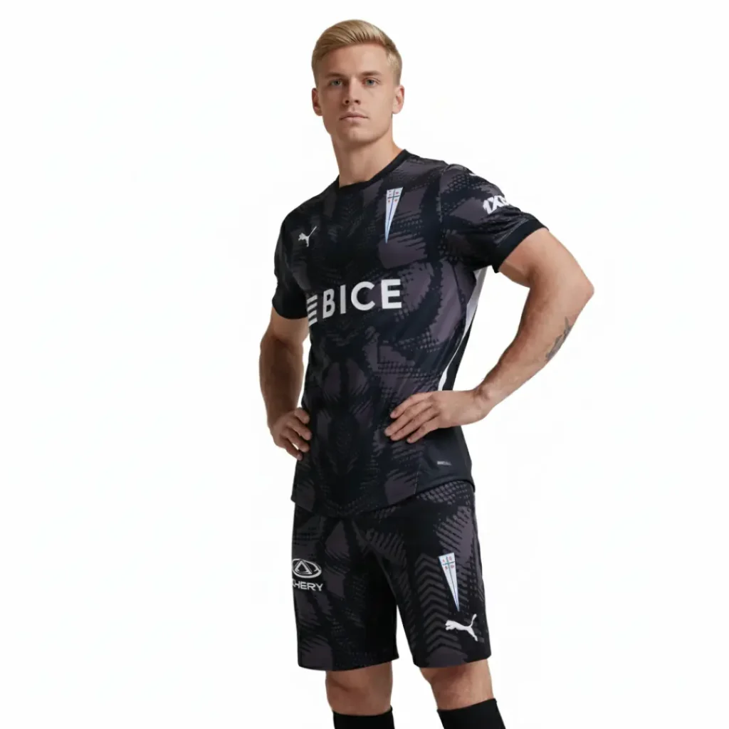 Hombre atlético posando con el uniforme completo de portero de Universidad Católica 2024, que incluye una camiseta y shorts negros con un patrón gráfico gris abstracto. La camiseta muestra los logos de BICE, Puma y CDUC, mientras que los shorts llevan los logos de Chery y Puma.